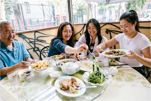 Bill, Judy, Kaitlin and Sarah from The Woks of Life sit at a round table with a lazy susan with Chinese food arrayed